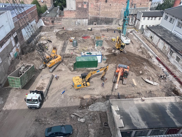 Plac budowy Ogrodowa 12 w Poznaniu z koparkami, maszyną do palowania i kontenerami budowlanymi, widok z góry.