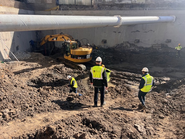 Koparka CAT oraz pracownicy w odblaskowych kamizelkach przy głębokim, zabezpieczonym rurami wykopie na placu budowy.