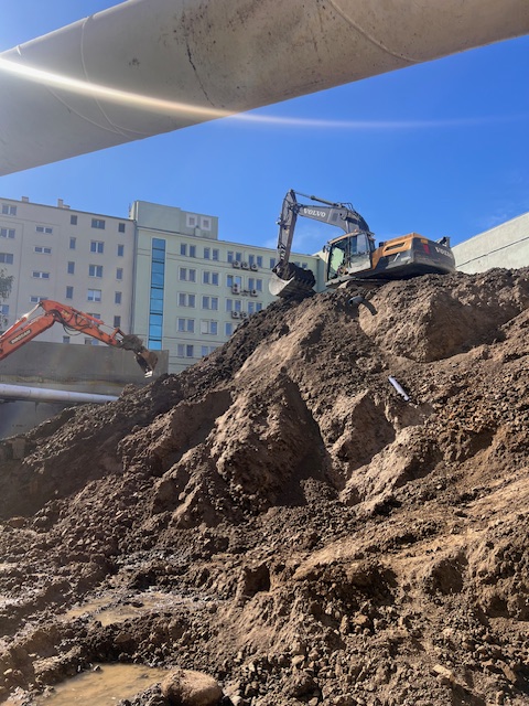Koparki na nasypie ziemnym podczas prac budowlanych przy zabezpieczonym rurami wykopie, na tle nowoczesnych budynków
