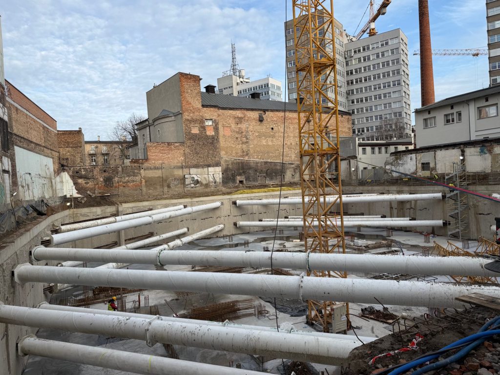 Plac budowy w centrum miasta z głębokim wykopem zabezpieczonym białymi rozporami, żółtym żurawiem wieżowym oraz otaczającymi go starymi kamienicami i nowoczesnymi budynkami.