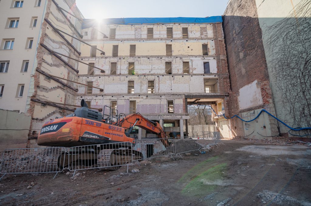 Zdjęcie przedstawia plac budowy inwestycji Ogrodowa 12 prowadzonej przez SOVO Development w Poznaniu. Widoczny jest fragment dawnego budynku, który przechodzi proces rewitalizacji oraz prac rozbiórkowych z użyciem koparki. Ściany budynku zostały zabezpieczone specjalnymi wspornikami, zapewniającymi stabilność podczas prowadzonych prac. Cały obszar budowy otoczony jest metalowymi barierkami ochronnymi, co podkreśla staranność i bezpieczeństwo realizowanych prac budowlanych. Pogodny dzień oraz nasłonecznienie podkreślają intensywny postęp prac przy projekcie.