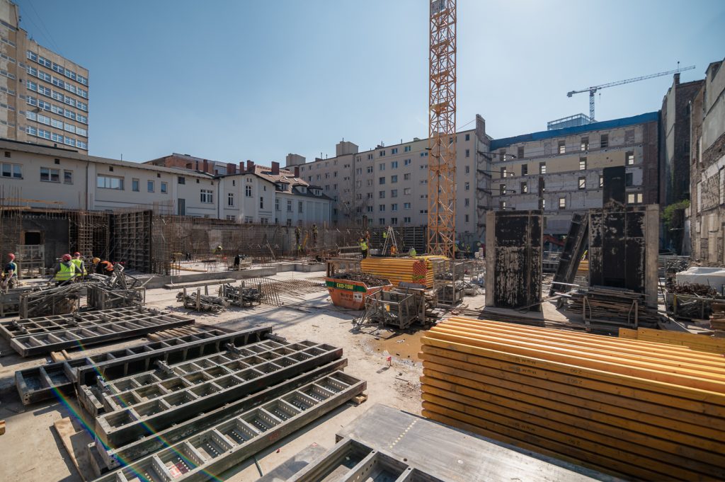 Zdjęcie przedstawiające aktualny stan prac budowlanych na inwestycji Ogrodowa 12 w Poznaniu, prowadzonej przez SOVO Development. Widoczny zakończony strop nad poziomem -1 oraz trwające prace nad konstrukcją nadziemną z udziałem dźwigu i ekipy budowlanej. W tle zabudowa centrum miasta.