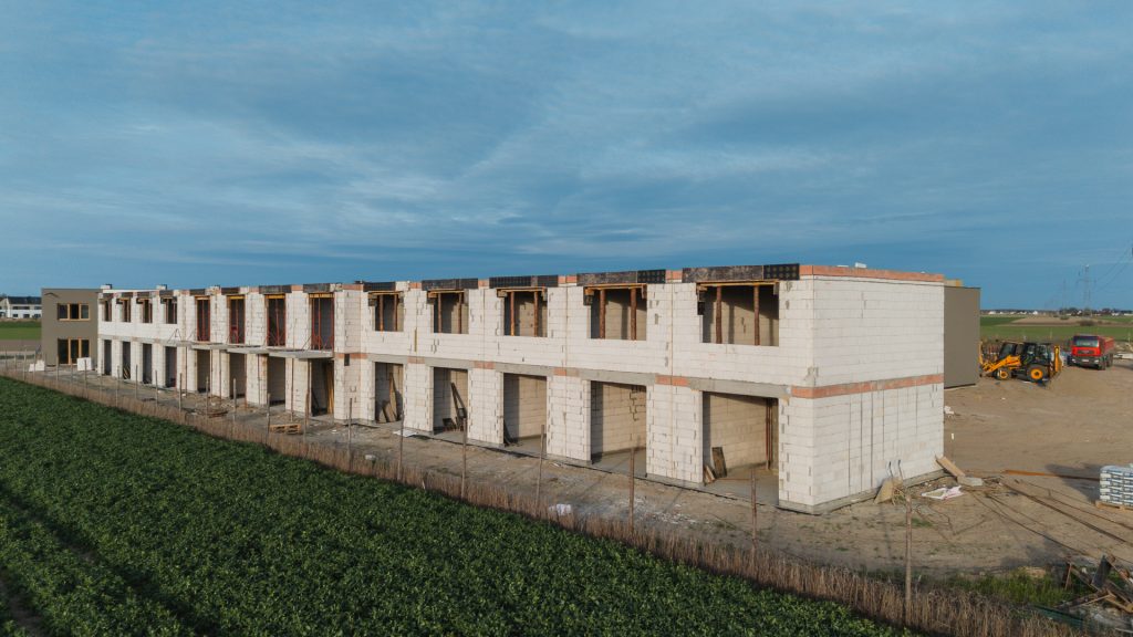 Budowa drugiego etapu inwestycji Rabowice Park w Swarzędzu – widok na szereg budynków w stanie surowym z zakończonym murowaniem drugiej kondygnacji, otoczenie rolnicze i sprzęt budowlany na placu.