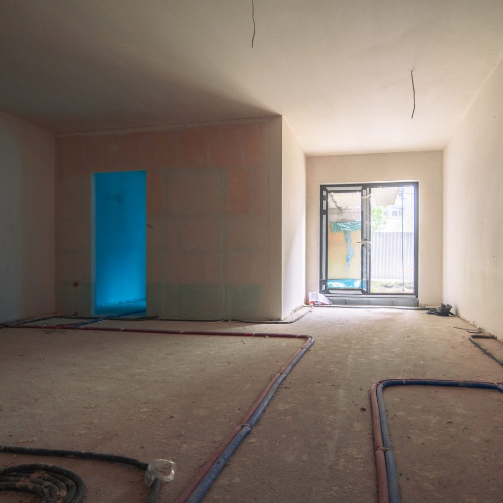 Wnętrze mieszkania w stanie deweloperskim z widocznymi instalacjami w podłodze i dużym oknem balkonowym.