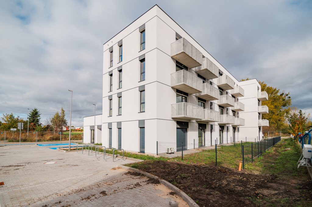 Nowoczesny biały budynek wielorodzinny z balkonami na osiedlu Tarasy Rokietnicy. Widok na gotową elewację i miejsca postojowe przed budynkiem. Inwestycja ogólnopolskiego dewelopera SOVO.