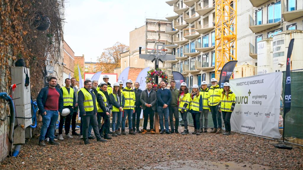 Uczestnicy uroczystości zawieszenia wiechy na inwestycji Ogrodowa 12, przedstawiciele generalnego wykonawcy i nadzoru budowlanego na tle powstającego budynku.
